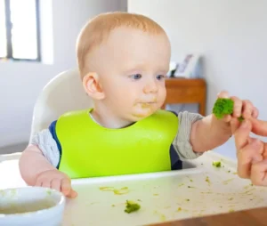 Smiling baby in a highchair, tasting pureed food with a spoon, symbolizing the start of solids